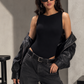 A woman modeling a black sleeveless bodysuit with a high neckline, styled with an open black leather jacket, dark gray wide-leg cargo jeans, and black sunglasses, standing against a textured gray concrete wall.