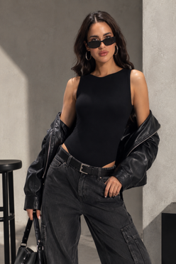 A woman modeling a black sleeveless bodysuit with a high neckline, styled with an open black leather jacket, dark gray wide-leg cargo jeans, and black sunglasses, standing against a textured gray concrete wall.